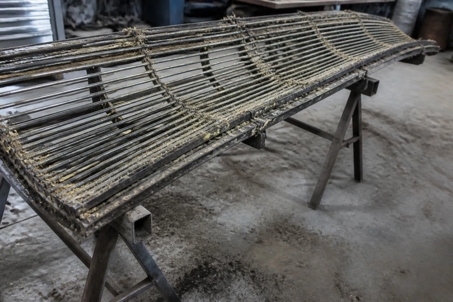 Un contre-batteur de moissonneuse déposé sur des tréteaux dans un atelier, grille de fils incurvée couverte de poussière de paille.