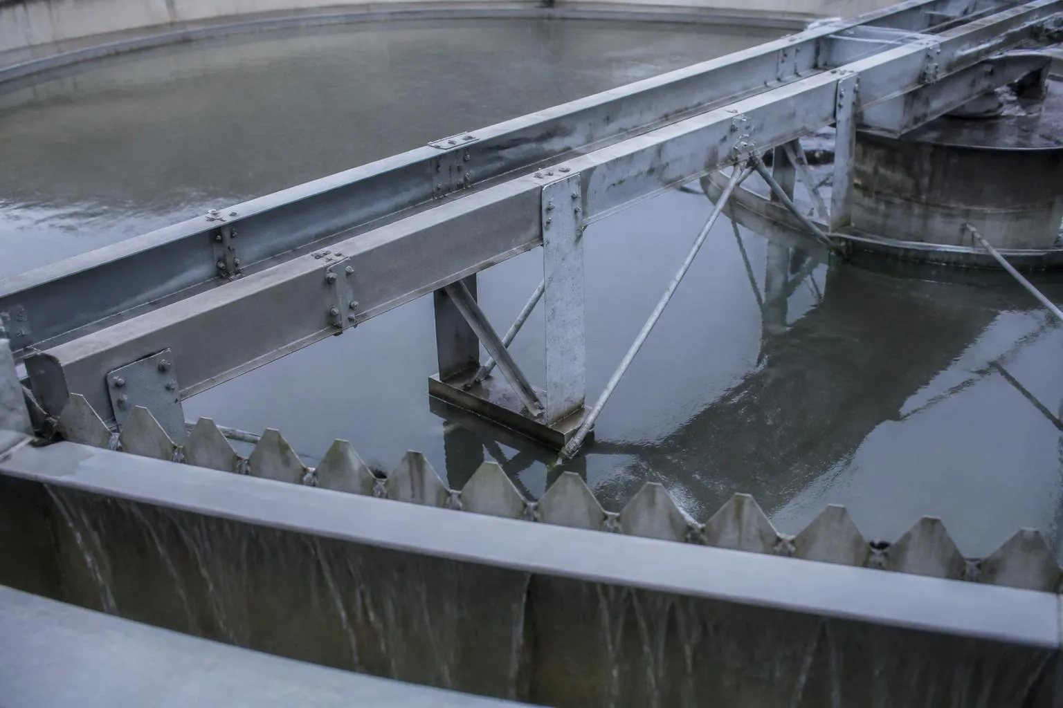 Le pont racleur d’un clarificateur circulaire au-dessus d’une eau grise, la lame déversante crénelée en bordure.